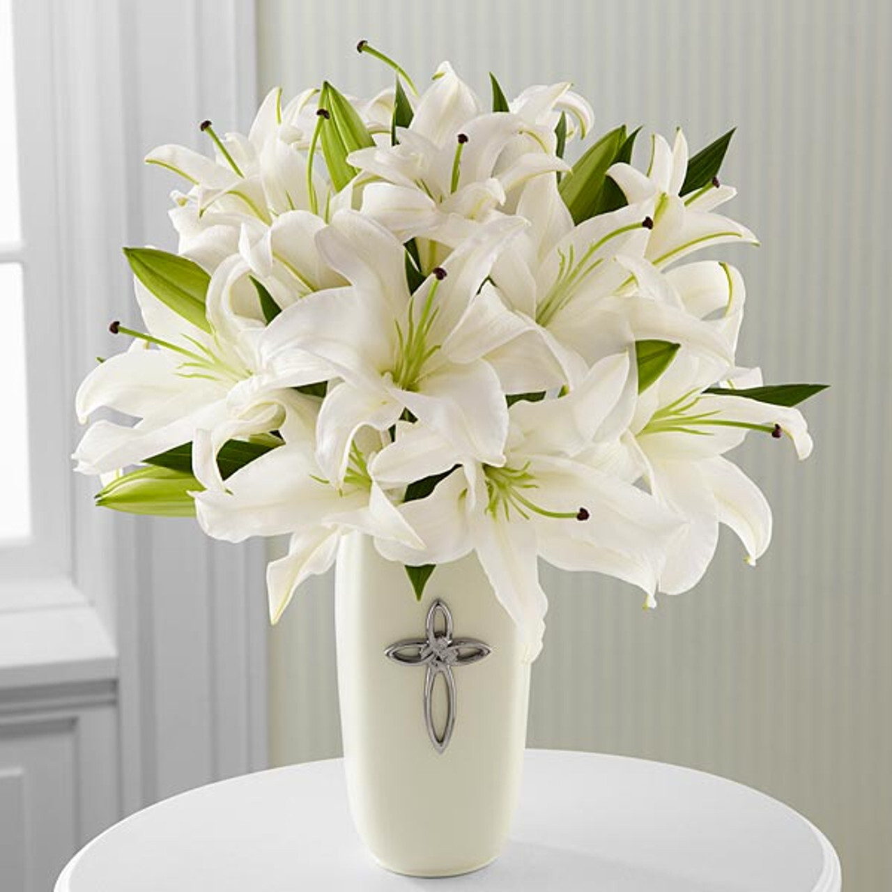 Elegant bouquet of white lilies with green leaves arranged in a white vase adorned with a silver cross.