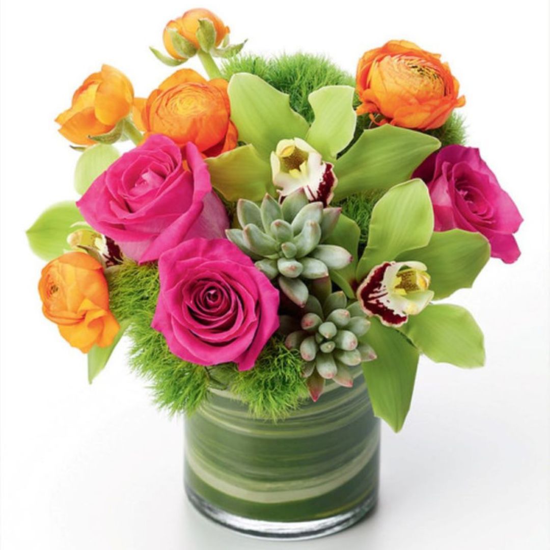 Colorful bouquet featuring pink roses, orange ranunculus, green orchids, and succulents in a clear vase.