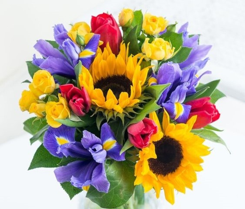 A colorful floral arrangement with vivid yellow sunflowers, purple irises, red tulips, and yellow roses in full bloom.