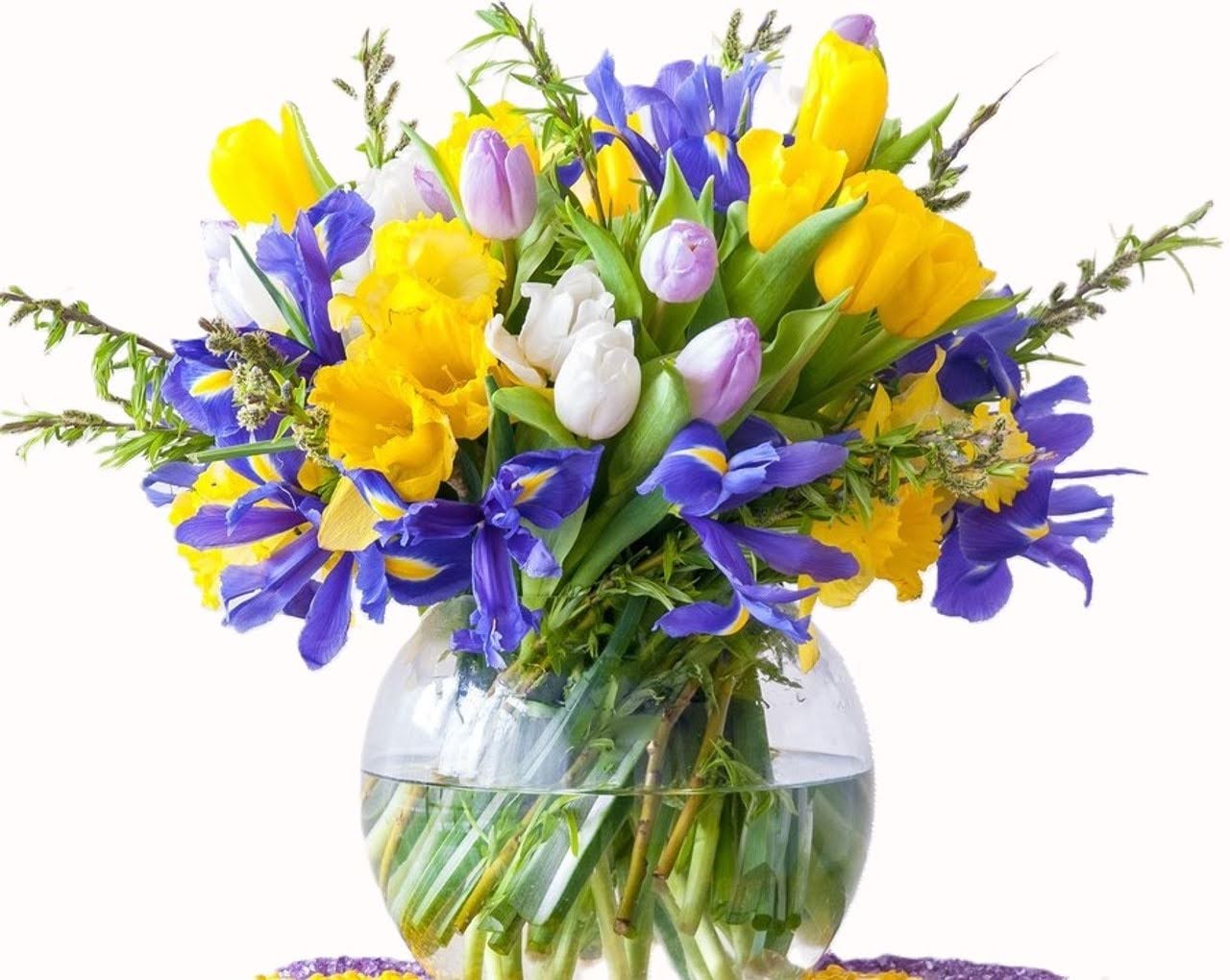Fresh purple iris, tulips, and yellow daffodils beautifully arranged in a round clear glass bowl.