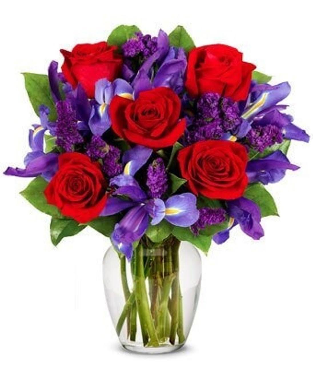 Vibrant bouquet with red roses, purple iris, and statice in a clear glass vase with green leaves.