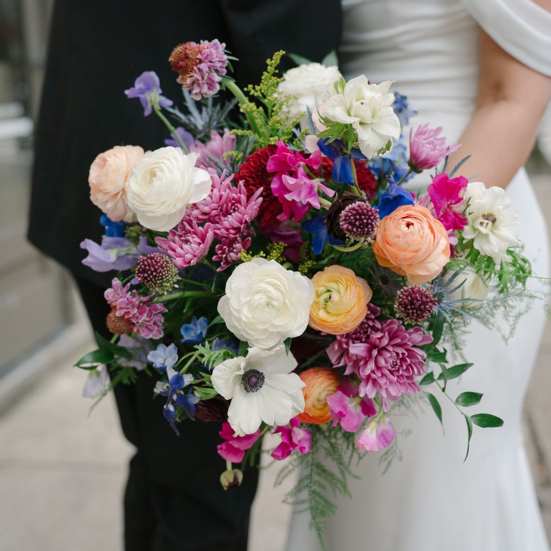 Close-up of a colorful wedding bouquet featuring ranunculus, anemones, and mixed blooms held by a bride in white.