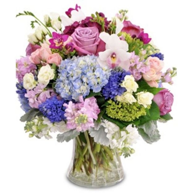 Mixed floral bouquet in a clear vase with lavender roses, blue hydrangeas, white orchids, and pastel filler blooms.
