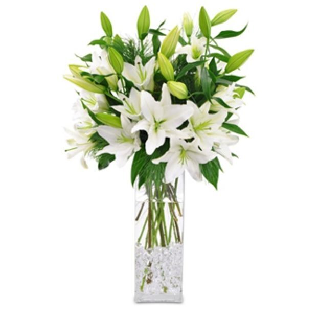 Tall glass vase filled with white lilies and green buds, arranged with leafy stems and clear decorative stones.