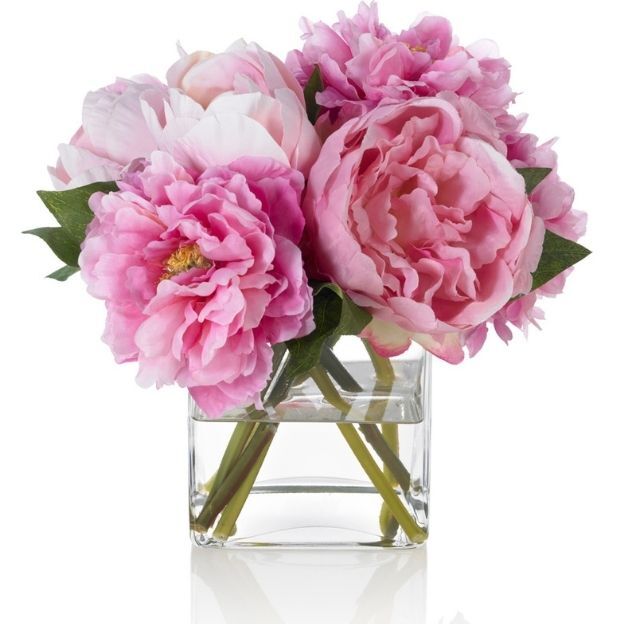 Pink peonies in full bloom arranged in a small, clear square glass vase with green stems visible.