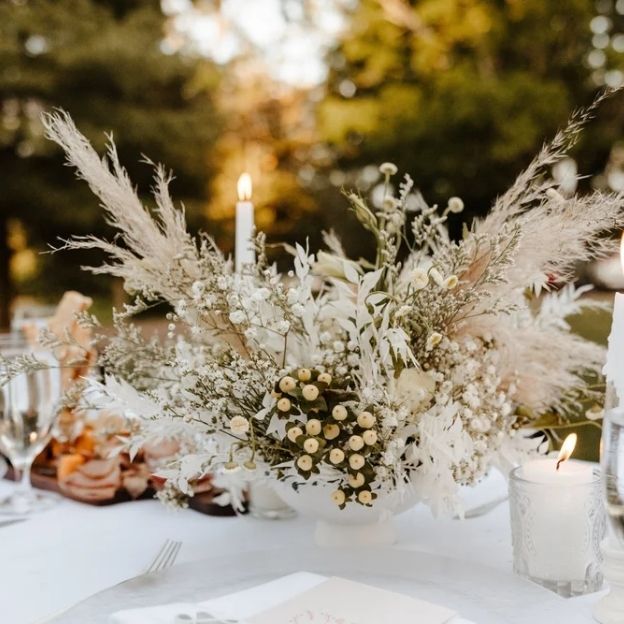 Neutral wedding centerpiece with pampas grass, white berries, and baby’s breath, flanked by glowing candles outdoors.