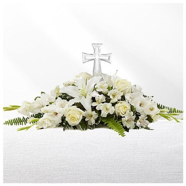 White floral arrangement with lilies, roses, and ferns, centered around a silver cross, displayed on a white cloth.