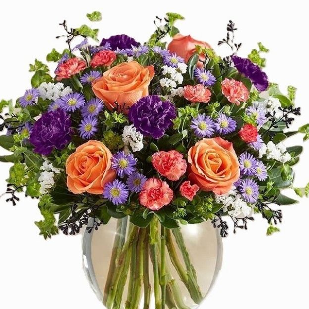 Mixed floral arrangement in a clear vase with orange roses, purple and pink blooms, white accents, and green foliage.