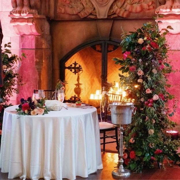Elegant round table set for two with floral decor, beside a fireplace and lush flower arrangement, champagne bucket nearby.