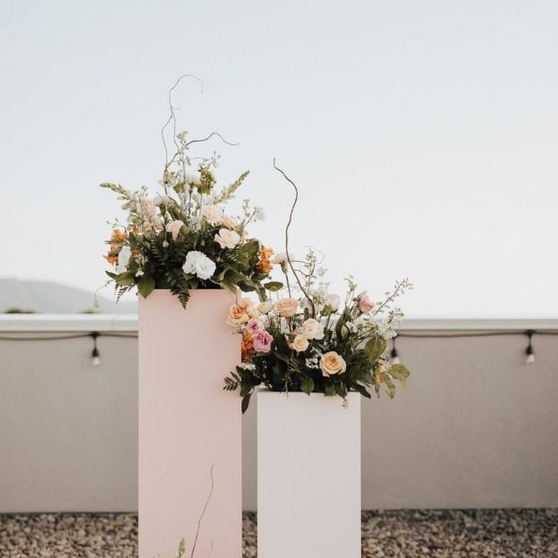 Two tall pastel pedestals with lush floral arrangements of roses and greenery against a clear sky backdrop.