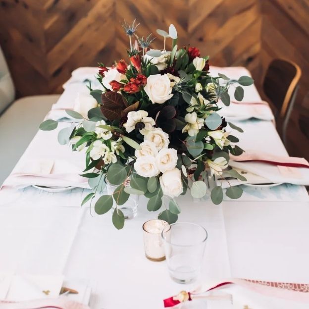Elegant table setting with white rose–eucalyptus arrangement, red accents, white linens, and a small glass votive.