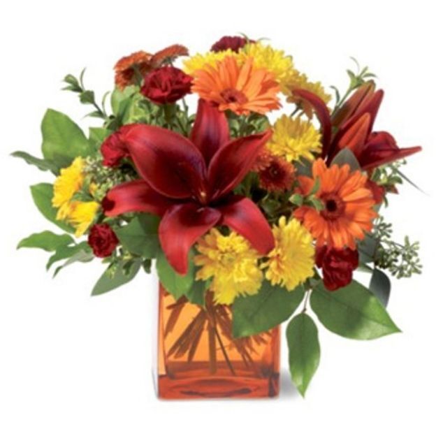 Autumn bouquet of dark red lilies, orange gerbera daisies, and yellow mums in an amber square glass vase.