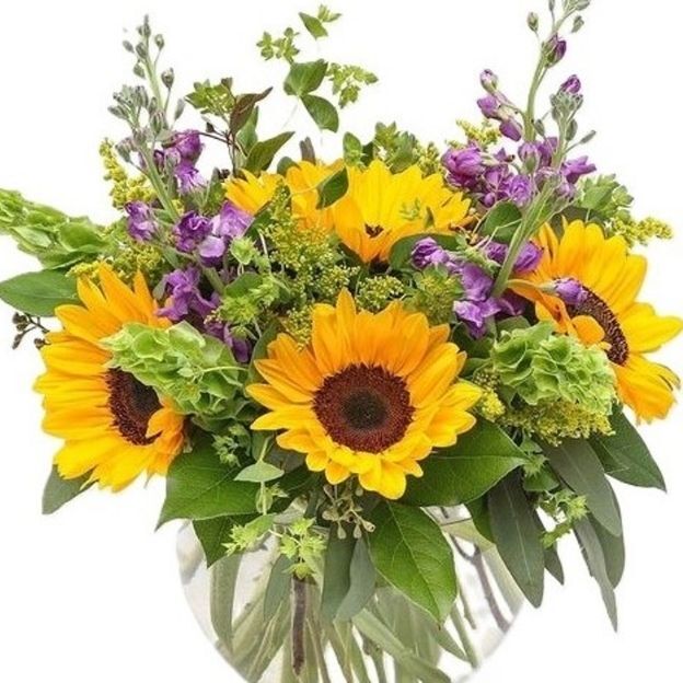 Bright sunflower bouquet with purple blooms and lime hydrangea, stems showing in a fishbowl glass vase.